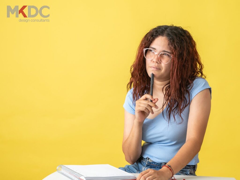 A woman with glasses, holding a pen, looking thoughtful against a yellow background.
